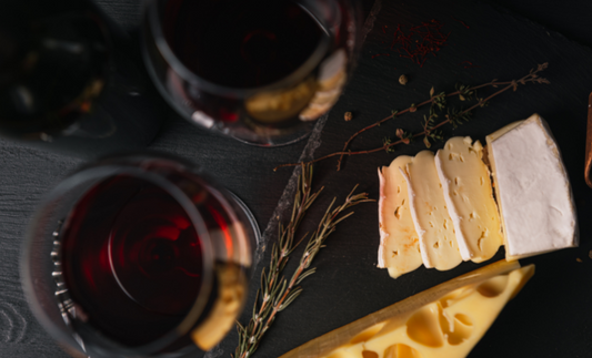 two glasses of red wine next to sliced cheese and herbs