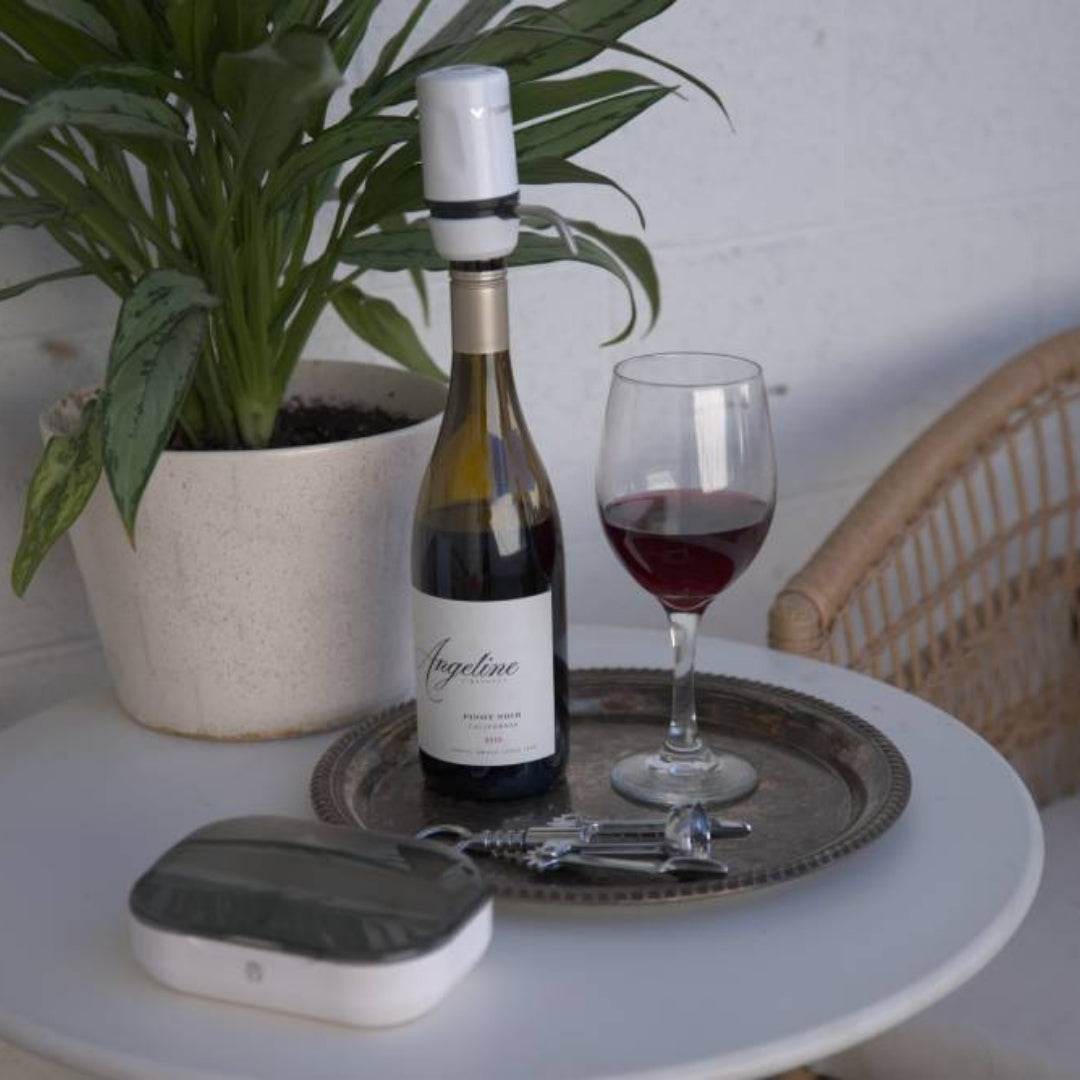 White Aervana Travel wine aerator on a red wine bottle next to a filled wine glass, a wine opener, and its carrying case