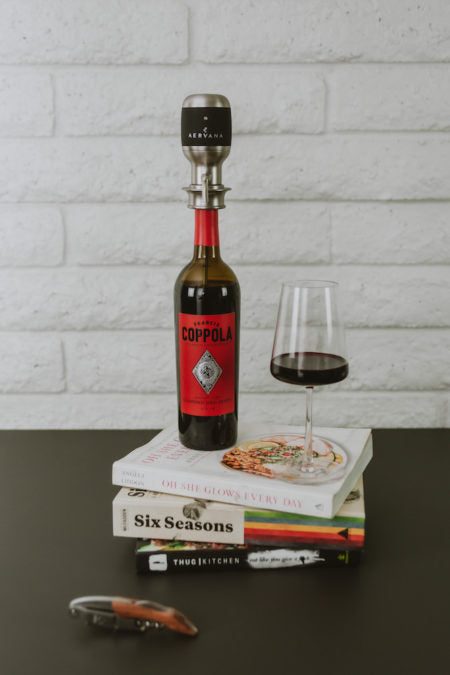 Original Wine Aerator on a wine bottle next to a filled wine glass sitting on top of a stack of books with branded corkscrew next to books
